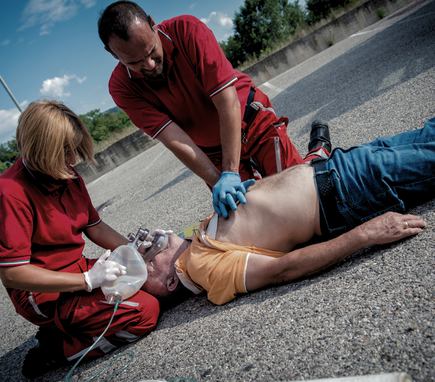 Two people in red uniforms providing first aid to a man lying on the ground, practicing chest compressions and oxygen mask use.