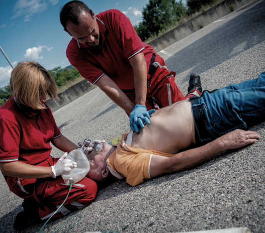 Two people in red uniforms providing first aid to a man lying on the ground, practicing chest compressions and oxygen mask use.